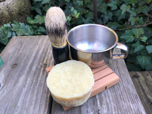 Shave brush, metal shave cup and off white round soap bar on a soap rack
